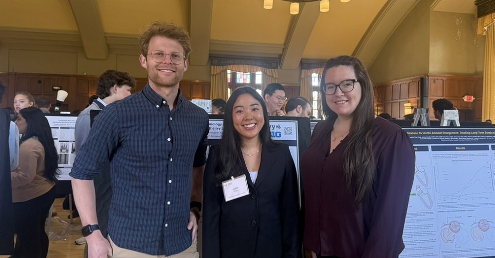 Three people wearing professional attire pose in front of a research poster during a research conference