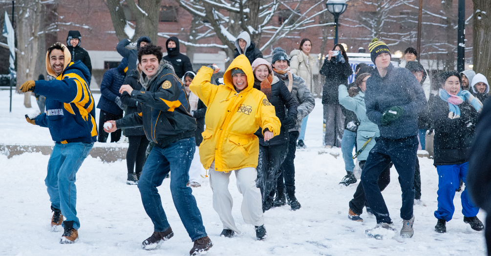 Students wearing University of Michigan winter gear engaging in a snowball fight at the Diag on campus on a snowy winter day