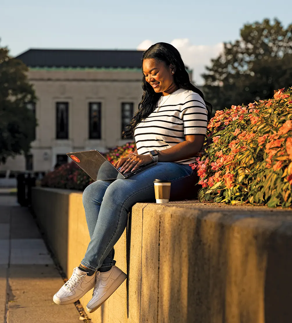 Jessica Holloway sits with her laptop on the edge of a bed of flowers on the Ann Arbor campus.