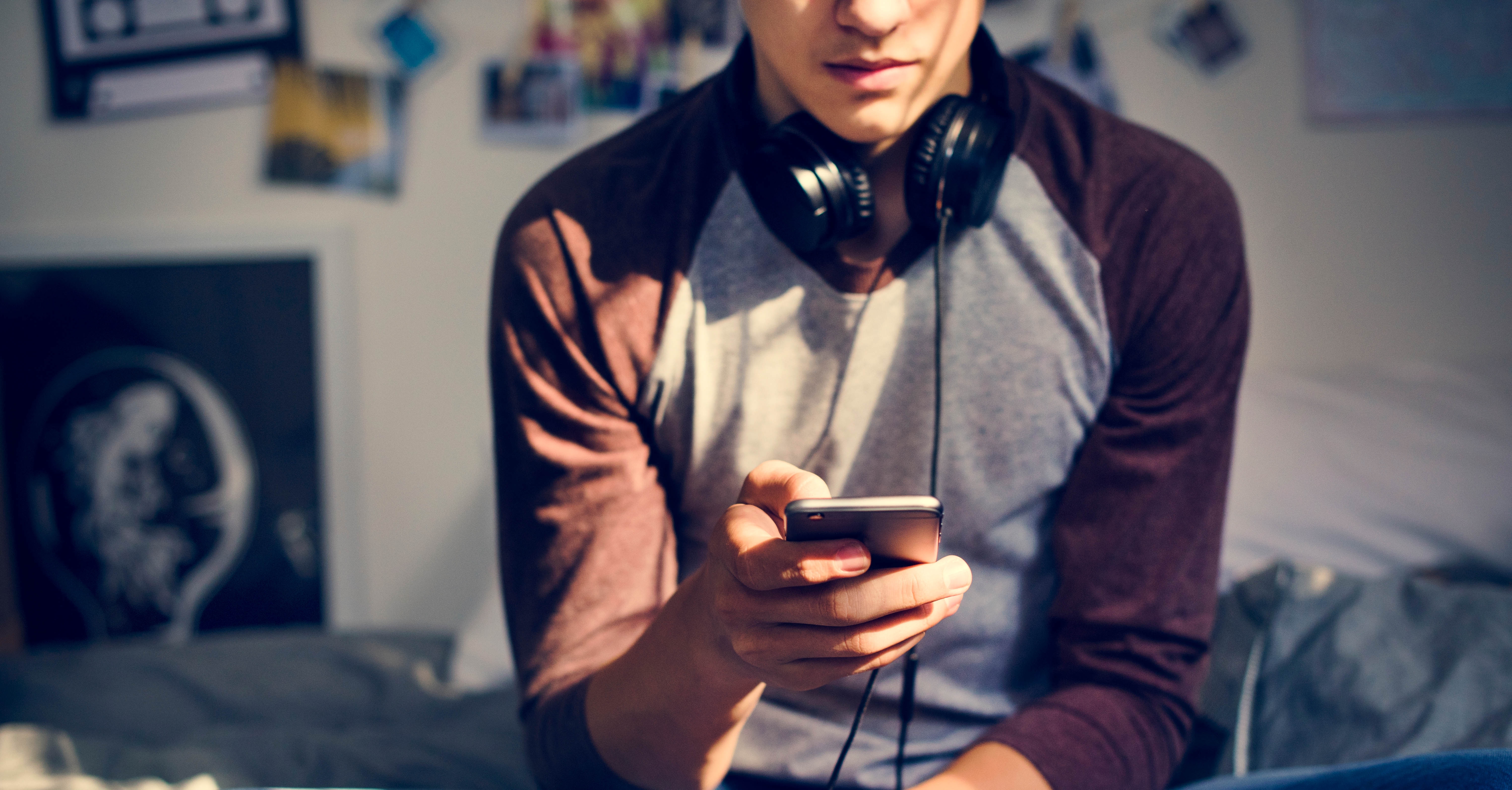 An adolescent male sitting on a bed looking at a cell phone.