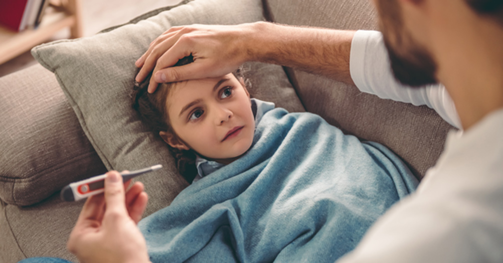 An adult checks the temperature on a thermometer while placing a hand on a young child's head. The child is wrapped in a blanket and laying on a couch. 