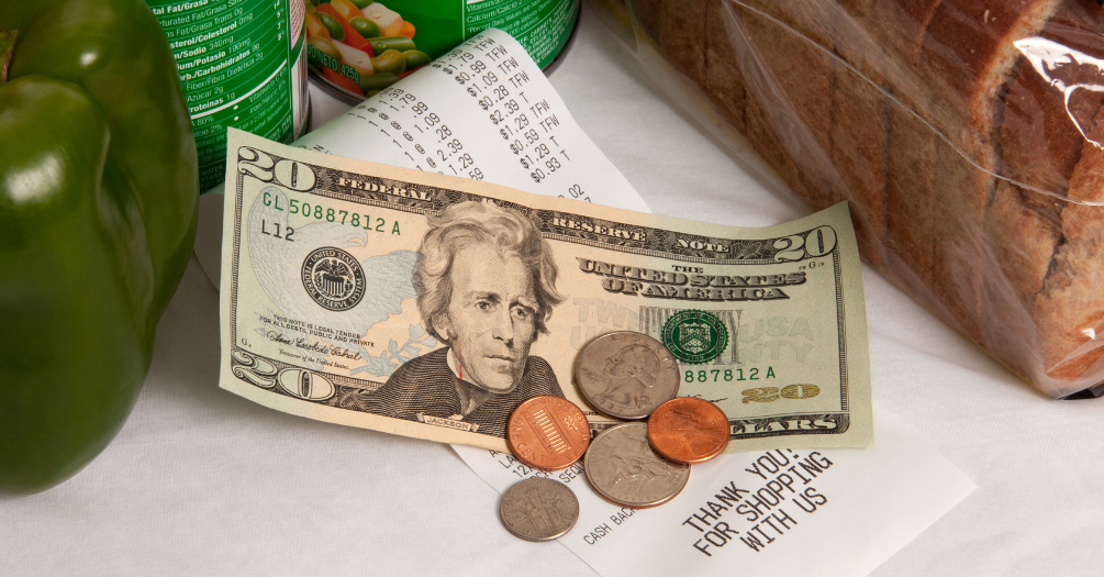 A twenty dollar bill and assorted coins lie on top of a receipt next to a bell pepper, canned vegetables, and a loaf of bread 