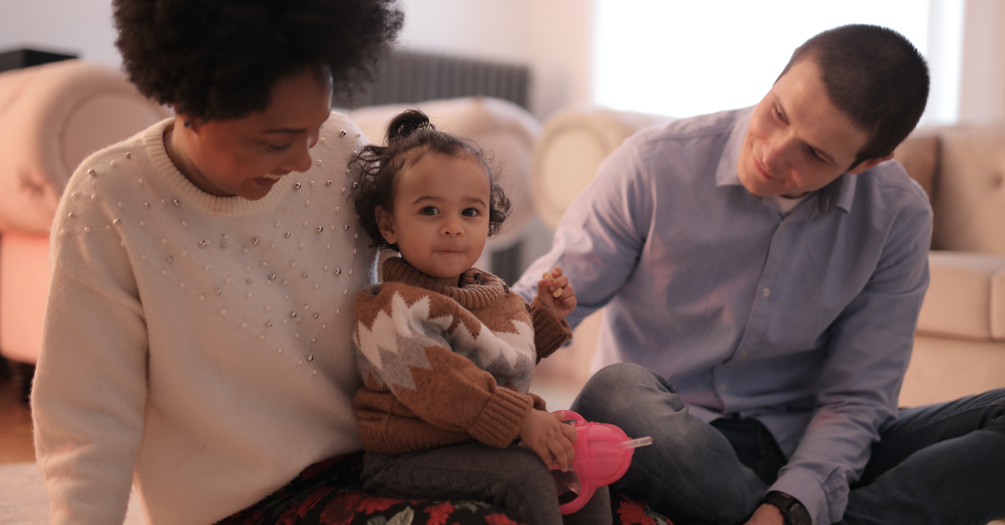 A mother and father are sitting on their living room carpet, while the mother holds a toddler on her lap.