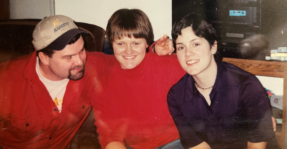 Three people sitting close together and smiling, with one wearing a cap labeled 