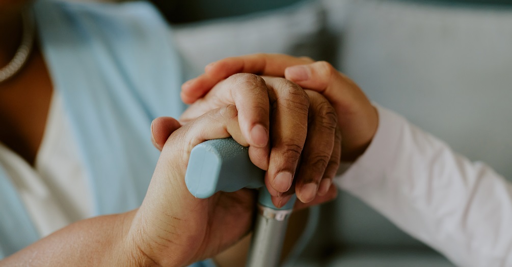 A child places their hand over an adult's hand who clutches a cane. 