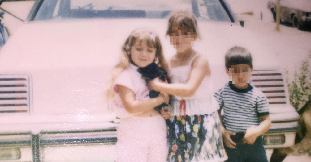 Three children posing in front of a white car. The child on the left is wearing a pink outfit and holding a black cat.